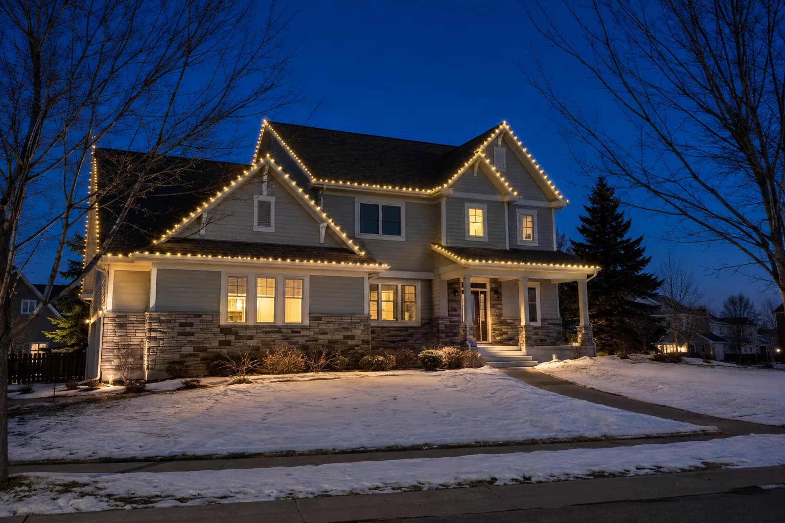 A two-story home at blue hour with warm white lighting tracing every gable and eave, snow on the lawn